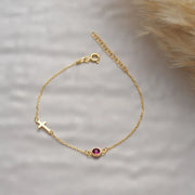 14k Gold side cross bracelet with a red birthstone on a soft gray background