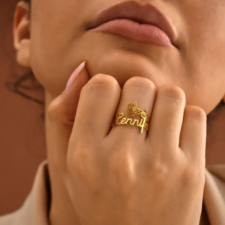 Close-up of a hand wearing a 14k gold filled birth flower name ring with 'Jennifer' on it, against a blurred background.