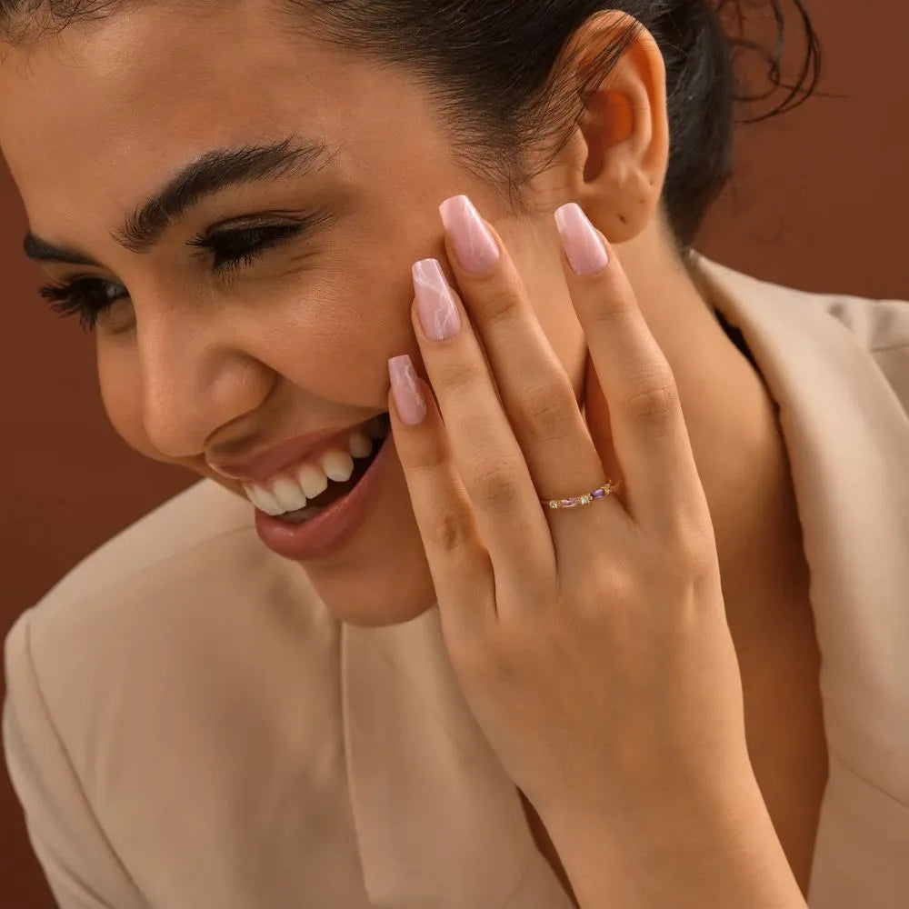 Woman with pink nail polish and a 14k gold-filled baguette birthstone ring on her finger, smiling against a warm-toned background
