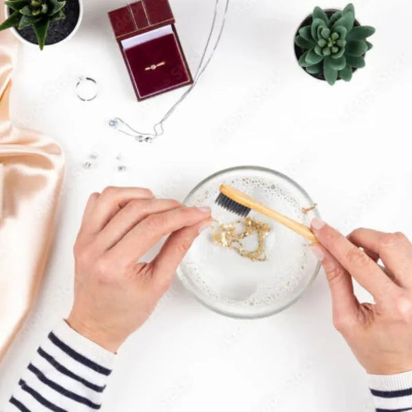 Person cleaning jewelry with a brush over a bowl of water on a white surface.
