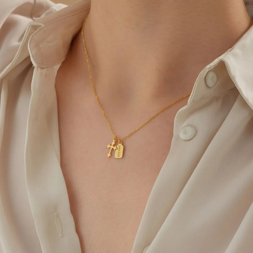 14k Gold necklace with two charms (cross and mini tag) worn by a person in a light-colored shirt.