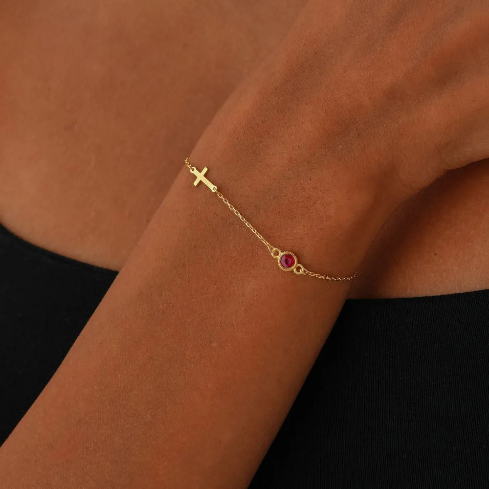 14k Gold cross bracelet with a red gemstone on a person's wrist against a brown background