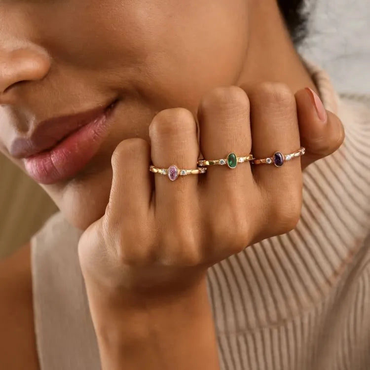 Close-up of a woman's hand wearing multiple birthstone cz diamond rings with gemstones. Gemstones: Pink tourmaline, emerald, and sapphire.