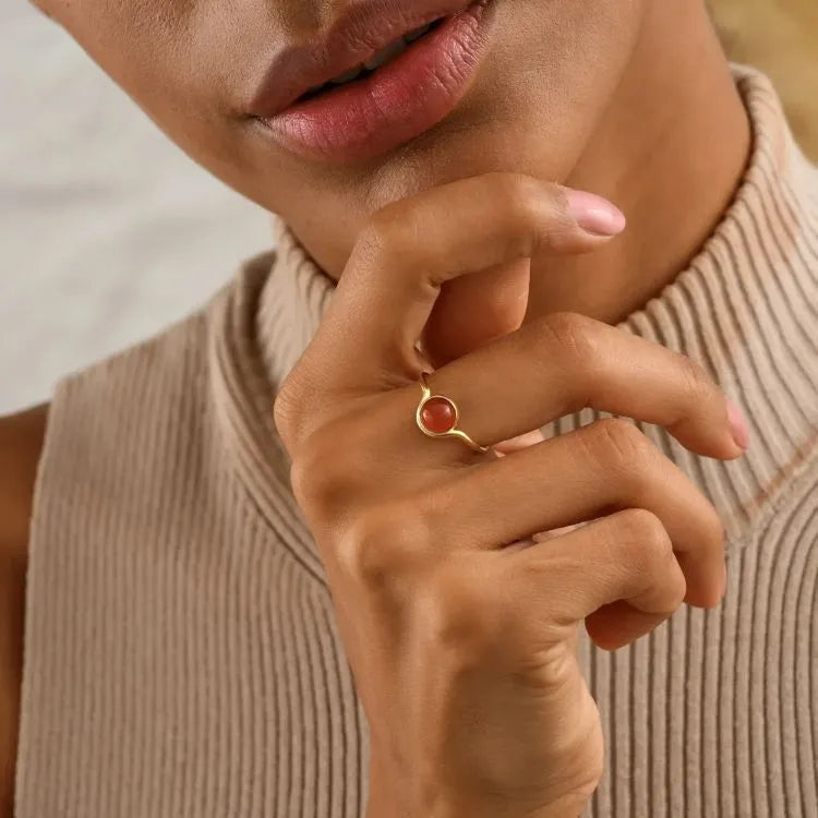 Close-up of a hand wearing a 14k gold filled ring with a red carnelian stone, against a neutral background.