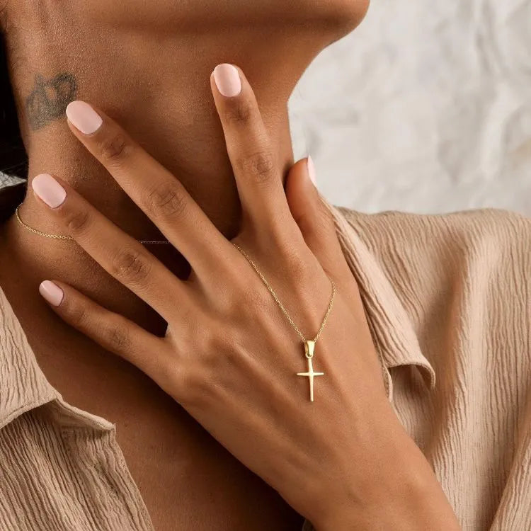 Woman hand wearing a 14k gold filled necklace with a cross pendant, with a neutral background.
