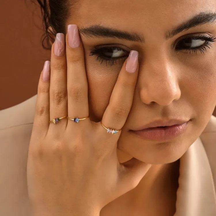 Close-up of a woman's face with one hand holding her cheek, wearing marquise birthstone rings.