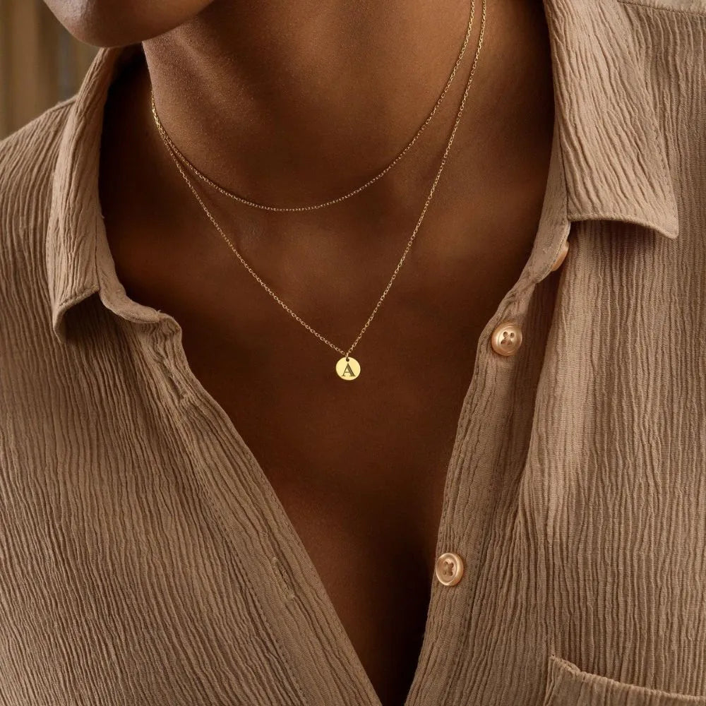 Close-up of a person wearing a 14k gold-filled tiny discs layer necklace with a small 'A' inital engraved pendant, set against a neutral background.