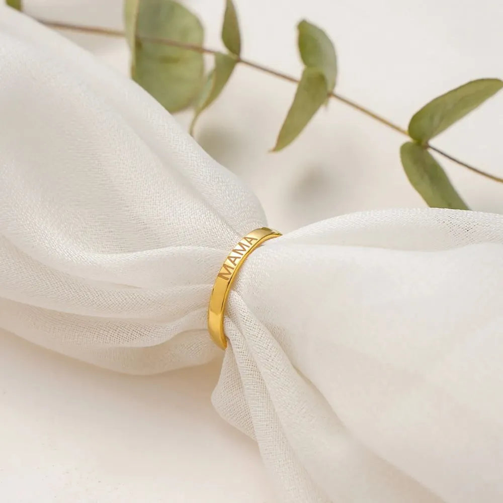 14k Gold-filled ring with 'MAMA' engraving on a white fabric background with green leaves.