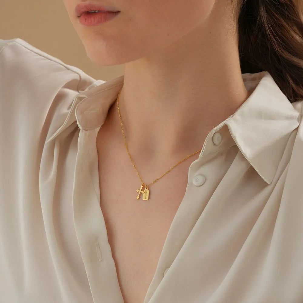 Close-up of a person wearing a 14k solid gold necklace with baroque cross and personalized mini tag against a neutral background