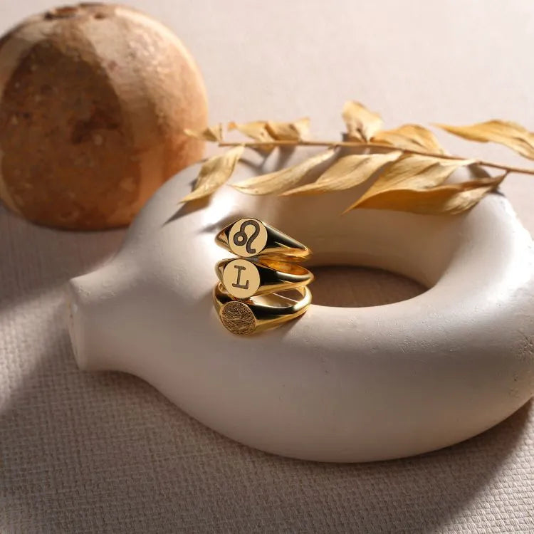 Three 14k gold filled rings with engraved initial, zodiac sign, and finger print on a white ceramic object with a natural background