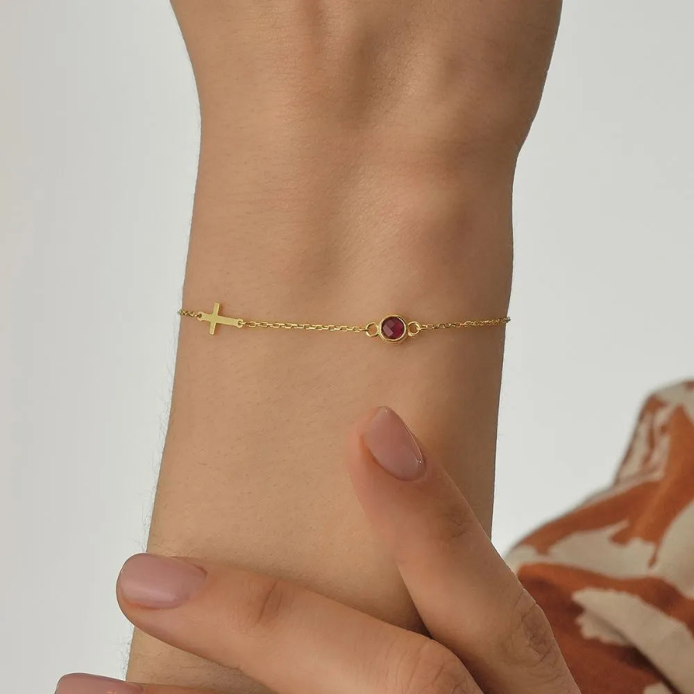 14k Gold cross bracelet with a red gemstone on a wrist against a neutral background