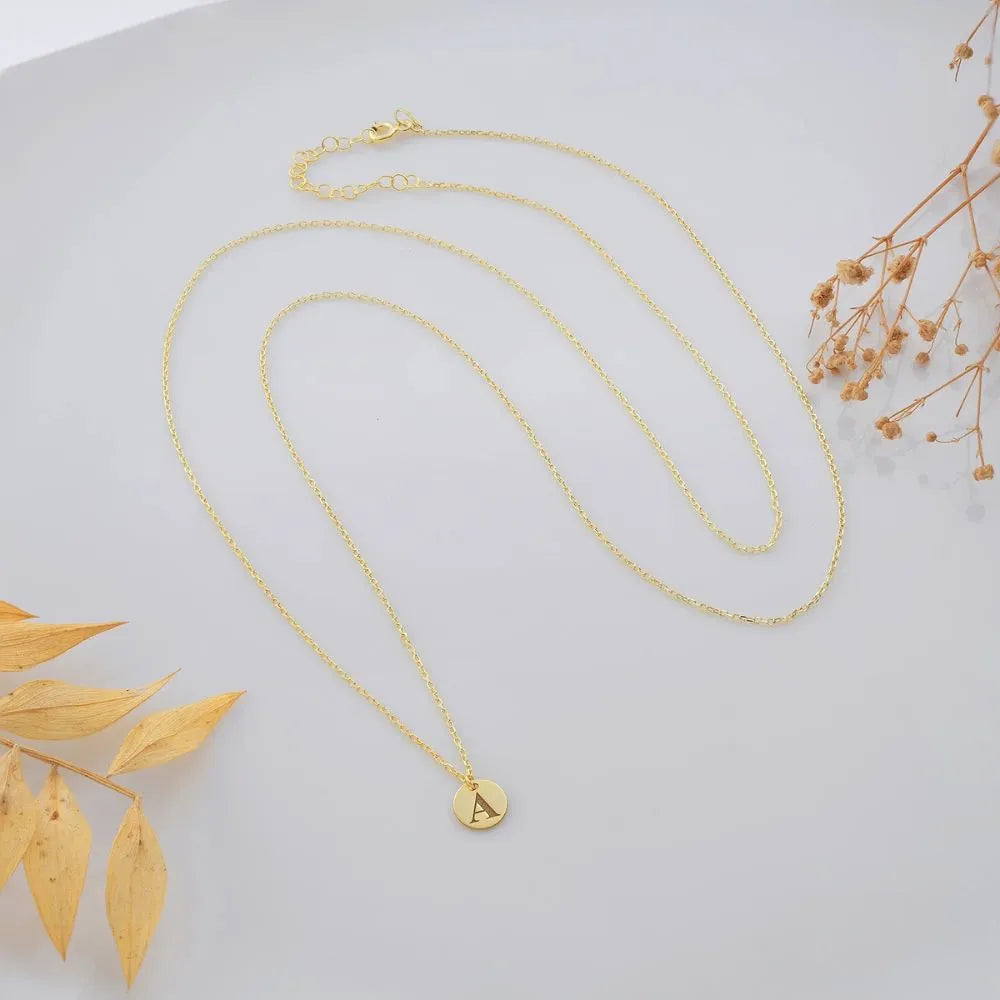 Tiny disc layered necklace featuring a round disc pendant engraved with the letter “A”, displayed on a light gray background with dried leaves and flowers.